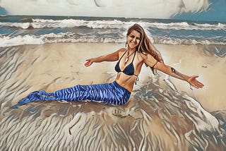 Woman at the beach in a blue bikini top and a blue fabric mermaid tail. Her is long and worn down and she’s wearing a shell necklace. She’s drawn to being a mermaid because doing it says something about myths, mermaids, and women’s power.