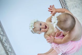 A baby dressed in a pink tutu and a flowery head-band delighting in her reflection in the mirror.