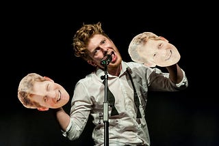 A photograph of an actor in a white shirt with a black bow tie hanging loose around his neck speaking into a microphone. He is holding two paper masks of Prince Harry’s face on his hands.