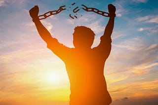 A shadowy photo of a man breaking his shackles with the sun and the sky in the background.