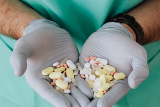 Medical professional wearing green scrubs and light blue latex gloves holding out two handfuls of a variety of pills.