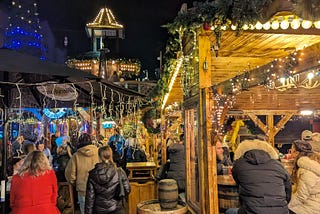Outdoor Christmas market beer garden at night, with wooden huts, warm fairy lights, people sitting at barrel tables, and a tall lit-up tree in the background.