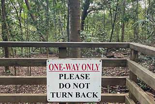 A sign nearing the end of the hiking trail reads in red and black bold, uppercase letters, “ One-way only” and “Please do not turn back.”