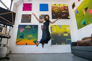 A woman in a long black dress jumps into the air in front of a wall on which 8 colorful geometric abstract paintings are hung.