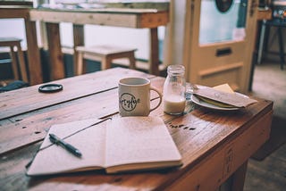 A notebook, a coffee mug and a little milk jug on a wooden table in a coffee shop.