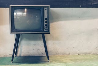 An CRT television on peg legs sits against a white industrial-looking wall.