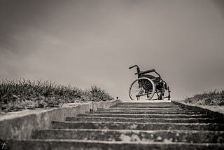 Black-and-white photograph of an empty wheelchair at the top of a stone staircase. The sky is overcast.