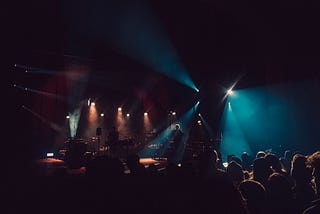 A dark photo of a crowd and a stage. A musician plays guitar as light washes over him.