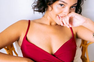 Lovely young woman in wine colored spaghetti-sleeved dress lounging in chair, head titled coquettishly back of right hand held against her mouth. She smiles slightly, seductively, in a come-hitherish way.