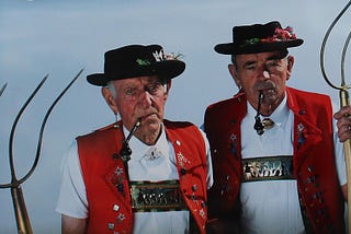 Two old and angry-looking Tyrolean farmers with red vests and pipes in their mouths, holding pitchforks.
