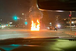 Road intersection with big flames in front of a Mini Cooper with debris scattered on the floor.