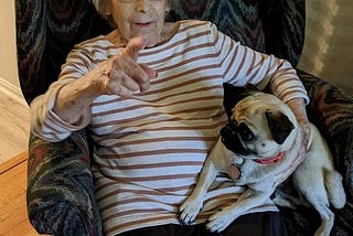 An elderly woman in a striped shirt and dark pants sits in an arm chair with a pattern in shades of green. She is pointing with her right hand and holding a young pug on her left side.