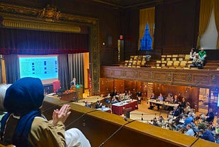Inside of Arlington Town Hall with people sitting above on the balcony and on the floor. A projector presents new bylaws for attendees to vote on. The room is full of residents listening in on debates and dialogue.