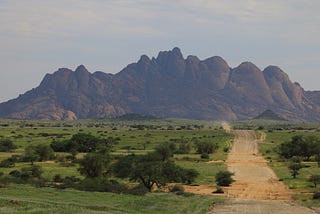 A gravel road leading between grassy meadows toward a granite mountain in the background.