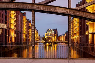 A gorgeous, twilight shot of buildings and other structures from the point of view of a person crossing over a canal, looking to one side; the water is in the center of the shot. Buildings are lit in gold.