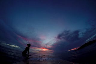 Sunset on a beach with dark blue skies and firey orange sun streaks, with a girl on her surfboard out on the line up