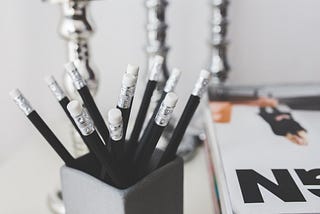 Nearly monochromatic photo of black pencils in a cup on a desk with other supplies on it.