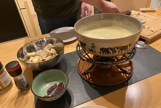 A pot of cheese sits on a burner on the table along with chopped bread and pieces of cured meats.