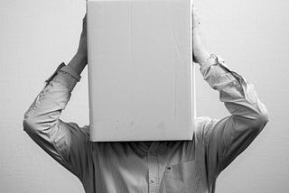 A black and white image of a man in a shirt holding a box that he has placed over his face and head