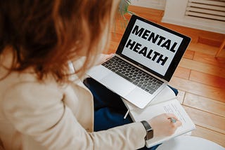 A therapist with long hair and a white jacket is balancing a laptop on their laptop while writing.