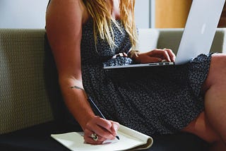 Tanned woman in black print dress sitting on couch with laptop in her lap and writing in a notebook on the couch.