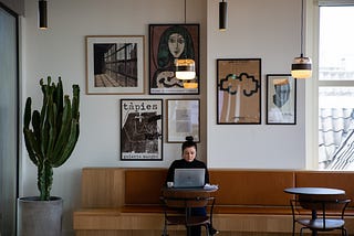 A woman sitting on a lounge area with a lot of frames on her background wall and working on her laptop