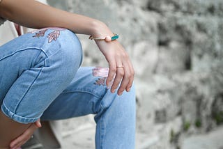 A young girl sitting down wearing jeans with pink flowers. She’s wearing a pink, gold and turquoise bracelet and a gold ring.