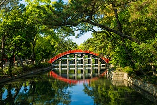 A photo showing green tree foliage over an arched red walking bridge reflected in the still water