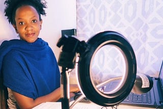 Woman sitting in front of her laptop. She is next to a camera light. She looking towards the camera and smiling.