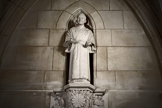 A carved limestone figure of Japanese pacifist Toyohiko Kagawa, originally sculpted by artist Frederick Hart. The figure wears an expression of understanding, his hands held forward, and partially open, as if in a gesture of giving. The figure is positioned in an arched niche in the center of the stone wall of the cathedral.