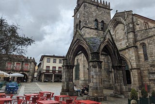 A medieval stone arch in front of a medieval church, with some red tables and chairs in front, under a dark cloudy sky.