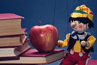 A string puppet stretching its arms toward an apple placed on a stack of books