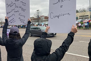 Two women dressed in black raincoats hold up white signs, one with “Silence = Compliance” and the other filled with a litany of issues (including but not limited to “Social Security,” “Education,” “Health Care,” “Research,” “History,” and “National Parks”), for passing cars on a wide avenue to see. Across the way, are other demonstrators with colorful handmade signs, including “RESIST.” (Photo by author.)