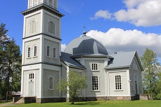 Colour photo from 2014 of Rautjärvi Lutheran church, Finland. A wooden church with a square wooden church tower topped by a metal-roofed spire. The church is painted light grey with white trim.