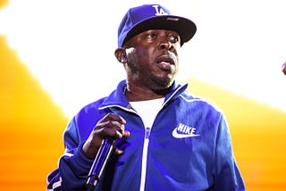 Rapper Phife Dawg holding a microphone on stage, wearing a blue baseball cap and matching track jacket
