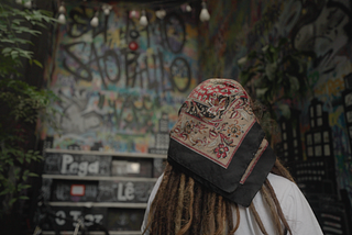 Foto de uma mulher de cabelos com dreads e lenço na cabeça. Ela está de costas para a câmera. Em frente a ela, está uma parede cheia de grafites.