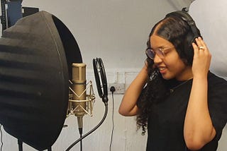 Malizah, standing next to recording kit, she is a oung Black woman with long black haid, and glasses. She is holding her headphones at each ear.