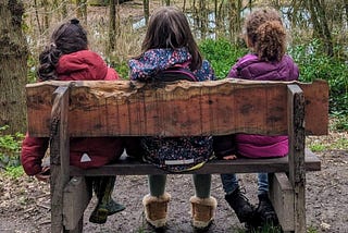 Three children sitting on a bench looking away from camera into a forest.