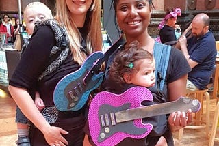 White woman and black woman carrying babies and foam guitars. Other people in the background