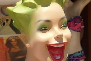 Head of mannequin with green hair and eye shadow, with red lips in a wide- open smile.