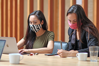 duas mulheres trabalham em uma mesa em ambiente corporativo usando máscaras