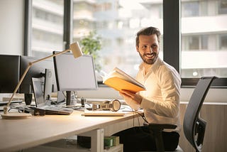 A man seated in front of a naturally lit office desk, smiling.