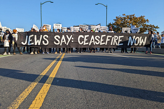 A road is completely blocked by a line of protesters. They’re holding a banner that says “JEWS SAY: CEASEFIRE NOW”