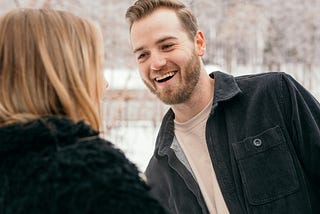 The image shows a woman from behind talking to a man who is facing her. We can see that he is making eye contact with her and looks like he may be chuckling at something she said.