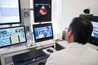 A medical doctor sits at a workstation with multiple monitors displaying a patient’s charts and a sonogram.