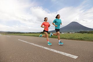 Two runners jog along a scenic road with a mountain in the background, wearing bright athletic gear and matching teal running shoes.