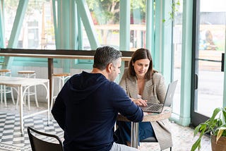 A man and a woman sit at a table and look together at the screen of a laptop.