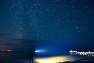 A person with a flashlight looking out over a dark ocean and the starry sky