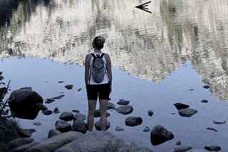 A girl standing in front of an alpine lake, her back to the camera, looking into the distance probably pondering who she is and the meaning of life