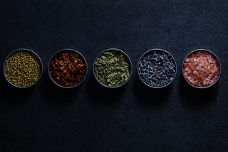 A dark textured background featuring five small round bowls arranged in a row, each containing a different culinary ingredient: coriander seeds, crushed red pepper, fennel seeds, lavender, and Himalayan pink salt.